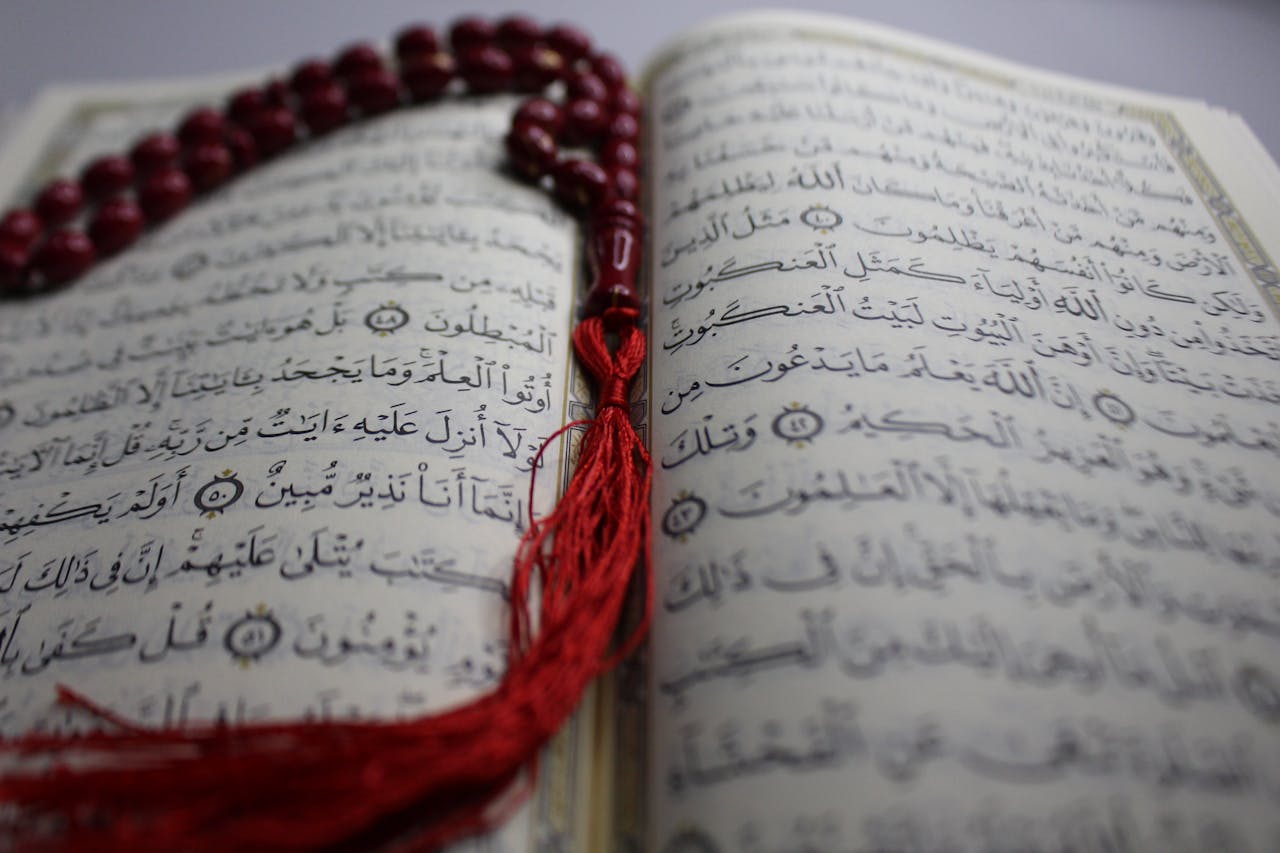 hero-img-01 Close-up view of an open Quran with red prayer beads, highlighting intricate Arabic script.